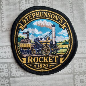 May include: A circular embroidered patch featuring a yellow and black steam locomotive on a blue sky background with clouds. The text STEPHENSON'S ROCKET 1829 is stitched in gold lettering on black banners at the top and bottom of the circular border.