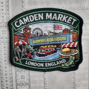 May include: A rectangular embroidered patch featuring the text CAMDEN MARKET and LONDON ENGLAND. The design depicts a clock tower, a Ferris wheel, a canal boat, a leather jacket, a vinyl record, a burger, and a green boot with a Union Jack flag.