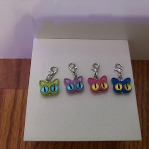 May include: Four metal cat-shaped charms with lobster claw clasps. The charms are colored lime green, purple, magenta, and blue, each featuring large, prominent eyes with yellow or blue pupils, designed for attachment to jewelry or accessories.