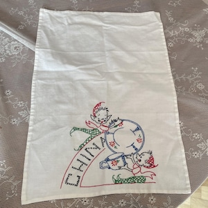 May include: A white rectangular kitchen towel featuring embroidered illustrations of two elves. One elf holds a plate and the other holds a teacup. The word DINNER is embroidered in black capital letters along a curved red line.
