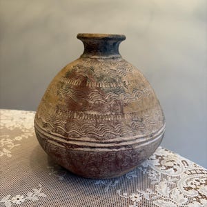 May include: A round, bulbous earthenware vessel with a narrow neck and a wide, textured body. The surface features alternating bands of incised wavy lines and horizontal grooves, finished in earthy tones of reddish-brown, tan, and dark gray.