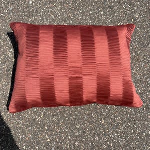 Op de afbeelding: Een rechthoekig sierkussen in een diepe terracotta oranje kleur. De stof heeft een opvallende verticale geplooide textuur die afwisselende banden van licht en schaduw creëert over het gehele oppervlak van het kussen.