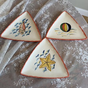Può includere: Tre piatti triangolari in ceramica con superfici bianco sporco e bordi arancioni testurizzati. Ogni piatto presenta un disegno dipinto a mano a tema spiaggia: una conchiglia, un pallone da spiaggia colorato e una stella marina gialla.