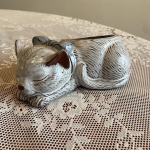 May include: A ceramic planter shaped like a sleeping cat with a glossy white and brown glazed finish. The cat has textured fur details, closed eyes, and a bow around its neck. The opening for plants is located on the back of the cat figure.
