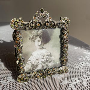 May include: A square metal picture frame with an ornate scrollwork border encrusted with small green, amber, and clear rhinestones. The frame holds a black and white portrait of a child and features the printed text size 3.5 inches x 3.5 inches.