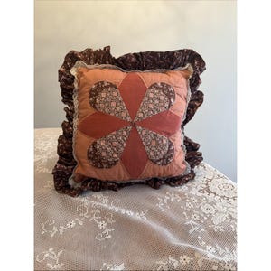 May include: A square throw pillow featuring a central six-petal flower applique in solid terracotta and brown floral fabric. The pillow is bordered by a white lace trim and a wide, gathered dark brown floral ruffle around the edges.