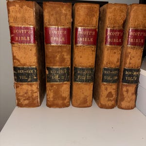 Puede incluir: Un conjunto de cinco libros antiguos titulados Scott's Bible, volúmenes I al V. Cada libro presenta una cubierta de cuero color canela desgastada con etiquetas rojas y negras en el lomo con letras doradas. Los lomos muestran los volúmenes.
