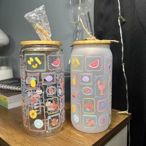 May include: Two cylindrical glass tumblers with bamboo lids and clear straws. One tumbler is clear glass, and the other is frosted glass. Both feature a repeating pattern of colorful postage stamp-style frames containing illustrations of food and sea life.