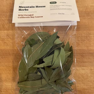 May include: A clear plastic bag filled with green California bay leaves, topped with a white label reading Mountain House Herbs, Wild Foraged California Bay Leaves, 0.5 oz, ~70 leaves, and Harvested 7/21/26. The label notes they are fresh-picked.