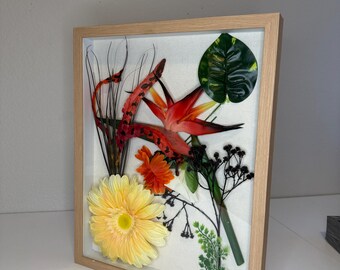May include: A square shadow box frame with a light wood finish containing a pressed floral arrangement. The display features a large yellow gerbera daisy, an orange flower, a red bird of paradise, a green monstera leaf, and thin dark stems.