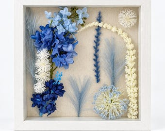 May include: A square white shadow box frame containing a dried floral arrangement on a beige linen background. The display features blue and navy hydrangea-like blossoms, light blue pampas grass, white spherical flowers, and a curved cream stem.