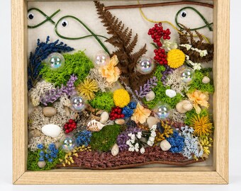 May include: A square wooden shadow box frame containing a nature-inspired collage. The display features green moss, dried ferns, colorful flowers, small white pebbles, and red berries. Two green wire fish shapes with googly eyes and iridescent bubbles.