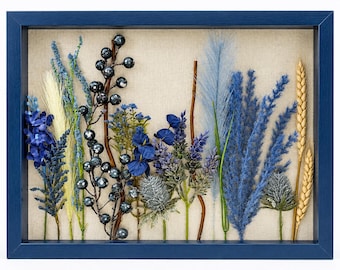 May include: A rectangular shadow box with a dark blue frame containing an arrangement of dried and artificial botanical elements. The display features blue berries, blue and cream grasses, blue flowers, thistle, and golden wheat stalks on linen.