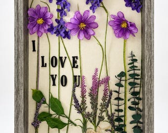 May include: A rectangular shadow box frame with a gray wood grain finish containing pressed purple flowers, green stems, and leaves on a cream background. The black text I LOVE YOU is arranged vertically and horizontally among the floral stems.