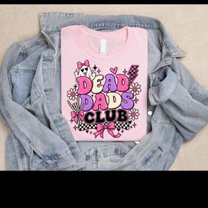 Pode incluir: Uma t-shirt rosa claro com uma estampa gráfica contendo o texto "DEAD DADS CLUB" em letras ousadas e coloridas. O design inclui um fantasma com um laço rosa, uma mão de esqueleto, flores, um raio e padrões xadrez em rosa e preto.