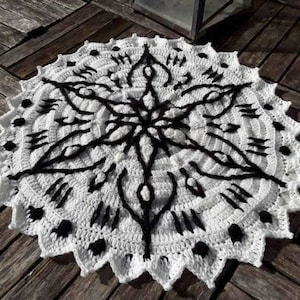 Può includere: Un tappeto rotondo in crochet bianco con un design geometrico nero. Il tappeto ha un bordo a festoni ed è realizzato con una varietà di punti.