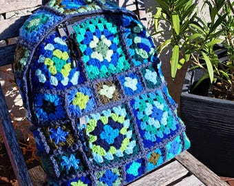 Op de afbeelding: Een handgemaakte gehaakte rugzak in tinten blauw, groen en bruin. De tas heeft een granny square design met een hoofdvak met ritssluiting en een zwart handvat. De rugzak ligt op een houten stoel.