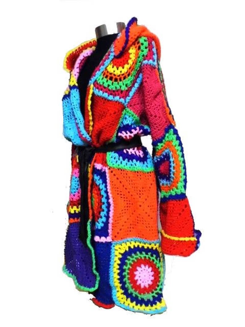 May include: A vibrant, handmade crochet coat in a patchwork design. The coat features a mix of bright colors including red, orange, blue, green, and pink. It has long sleeves and a hood, with a black belt cinching the waist. The coat is displayed on a mannequin.