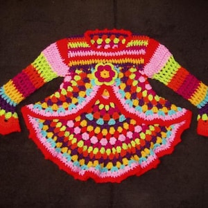 May include: A colorful crocheted baby dress with long sleeves and a bell-shaped skirt. The dress is made with a variety of bright colors, including red, orange, yellow, green, blue, and purple.