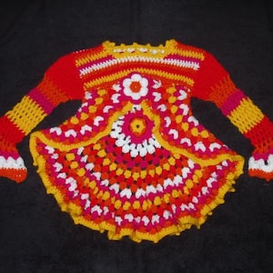 Könnte beinhalten: Handgefertigtes Häkeltop in leuchtenden Farben: Rot, Orange, Gelb und Weiß. Das Oberteil hat ein zentrales Blumenmuster und Glockenärmel. Die Häkelarbeit ist detailliert, mit einem gewellten Rand an Ärmeln und Saum. Ein farbenfrohes Kleidungsstück.