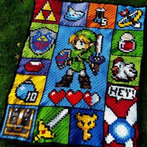以下が含まれることがあります： ゼルダの伝説のビデオゲームのリンクのピクセルデザインが施された、かぎ針編みの毛布。毛布は、剣、ハート、爆弾、鶏など、さまざまな色と画像の正方形で構成されています。