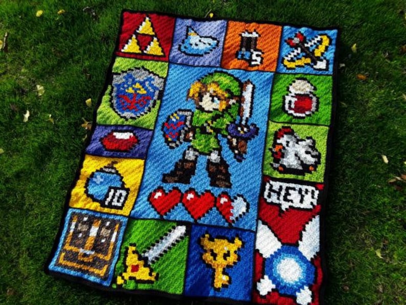 以下が含まれることがあります： ゼルダの伝説のビデオゲームのリンクのピクセルデザインを施した、かぎ針編みの毛布。毛布は緑、青、赤、黄、白の糸で作られています。デザインには、リンクの剣、盾、ハートが含まれています。