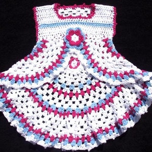 Könnte beinhalten: Weißes Häkelkleid für Babys mit rosa und blauen Akzenten. Das Kleid hat einen gewellten Saum und ein Blumenmuster auf dem Oberteil.
