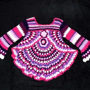 Könnte beinhalten: Ein gehäkelter Babysweater mit langen Ärmeln und einem glockenförmigen Saum. Der Pullover ist in verschiedenen Schattierungen von Rosa, Lila und Weiß gearbeitet.