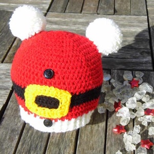 Peut inclure: Chapeau de Noël en crochet rouge et blanc avec une ceinture noire et une boucle jaune. Le chapeau a deux pompons blancs sur le dessus.