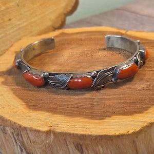May include: A silver cuff bracelet featuring a series of smooth, oval-shaped orange stones set in metal bezels. The band is decorated with alternating metal leaf motifs between each stone, creating a continuous pattern around the wrist piece.