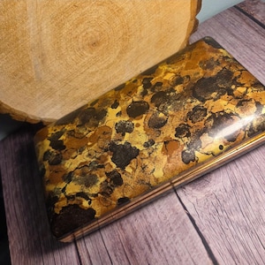 May include: A rectangular box featuring a glossy finish with a mottled pattern of dark brown, black, and metallic gold tones. The lid has a smooth, reflective surface with a distinct abstract design, while the base shows layered wood grain edges.