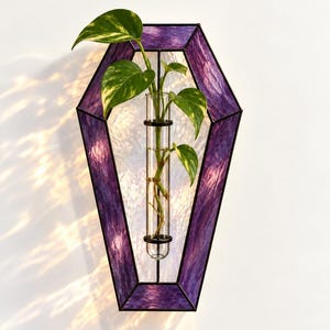 May include: A coffin-shaped wall planter made of purple and clear textured stained glass with black metal framing. A clear glass test tube is mounted in the center, holding a green and yellow variegated pothos plant cutting in water.
