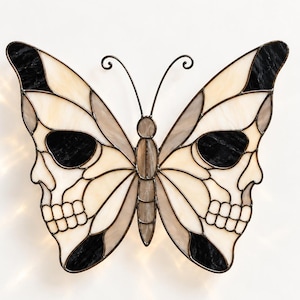 May include: A stained glass butterfly wall decoration featuring a cream and black color palette. The wings are designed with integrated skull silhouettes, outlined in dark metal solder, with two thin metal antennae at the top center.