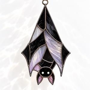 May include: A stained glass bat hanging ornament featuring black and iridescent light purple glass panels held together by dark metal solder lines. The bat is depicted in a hanging position with wings wrapped around its body and two white eyes.