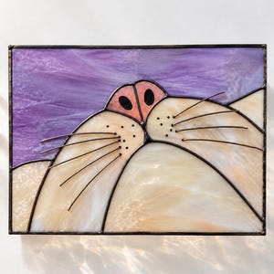 May include: A rectangular stained glass panel featuring a close-up of a cat nose and muzzle. The piece uses cream-coloured glass for the muzzle, a pink glass nose with black nostrils, and a purple background. Metal wire whiskers are attached.