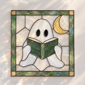 Puede incluir: Un panel de vidriera cuadrado que muestra un fantasma blanco con ojos ovalados negros leyendo un libro verde. Una luna creciente amarilla se sitúa en la esquina superior derecha. La pieza está enmarcada por un borde de vidrio verde y crema.