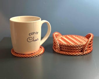 May include: A cream-colored ceramic mug with the text Cup of Cheer printed in black, resting on a round woven brown coaster. Beside it sits a matching stack of round woven brown coasters held in a small, circular woven basket holder.