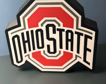 May include: A black, red, white, and gray Ohio State logo sign. The design features the word OHIO STATE in bold black lettering centered over a red and white block shape with a thick black border and a textured finish on the front surface.