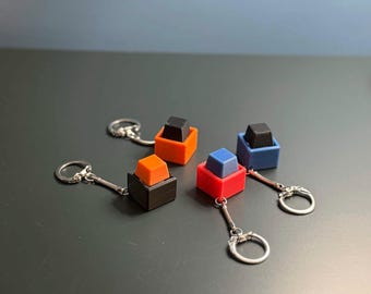 May include: Four mechanical keyboard switch keychains featuring square bases and contrasting colored keycaps. The set includes a black base with an orange cap, an orange base with a black cap, a red base with a blue cap, and a blue base with a black cap.