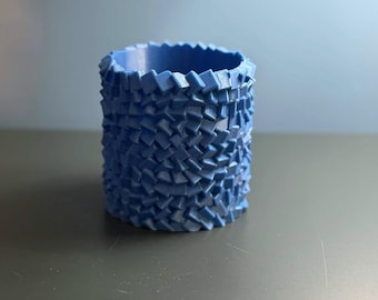 May include: A cylindrical blue container featuring a textured exterior surface composed of numerous small, repeating, angled rectangular blocks arranged in a dense, interlocking, brick-like pattern around the entire circumference.