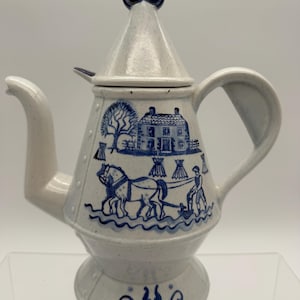 May include: A speckled off-white ceramic teapot with a conical lid and a dark blue finial. The body features a blue transferware-style illustration of a person plowing a field with a horse, a house, and trees. The base has a blue scroll design.