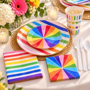 May include: A set of party supplies featuring rainbow-colored designs, including a large plate with diagonal stripes, a smaller plate with a radial sunburst pattern, a stack of striped napkins, a radial patterned napkin, and a paper cup.
