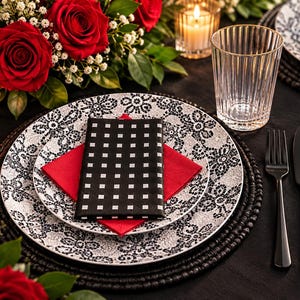 May include: A place setting featuring a white plate with a black floral lace pattern, topped with a red square napkin and a black napkin with a white grid pattern. A clear ribbed glass, black fork, knife, and spoon are arranged on a black tablecloth.