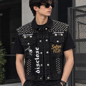 May include: A black denim vest featuring silver metal studs on the shoulders and front panels. The vest includes white text reading "Blackgoat" and "disclose," along with a skull graphic and yellow embroidered text reading "SHADOW OF FEAR."