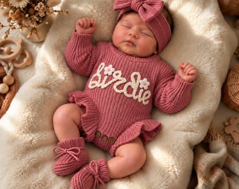 May include: A dusty rose knitted long-sleeve romper featuring a ruffled hem, three wooden buttons at the crotch, and the word "birdie" in white cursive text with two small white flowers. Includes a matching bow headband and knitted booties.