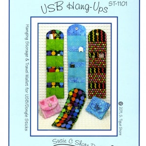May include: A sewing pattern for a USB hang-up storage and travel wallet. The pattern includes three different designs: a green and black design with peace signs, a blue design with white polka dots, and a black and white design with zigzags. The pattern also includes a small pink wallet with hearts and a blue wallet with a crown.