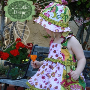 May include: A young girl wearing a pink and green floral dress with a matching sun hat. The hat has a green and white floral pattern with a green ruffle trim. The dress has a green and white polka dot ruffle trim. The girl is holding a small orange flower.