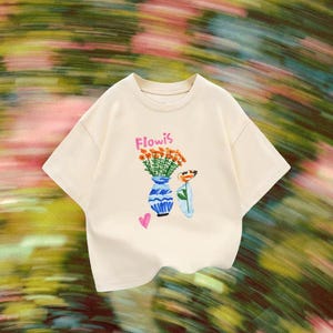 May include: A cream-coloured short-sleeve t-shirt featuring a graphic print of orange flowers in a blue and white patterned vase, a single flower in a clear vase, and a small pink heart. The text "Flowis" is printed in pink above the flowers.