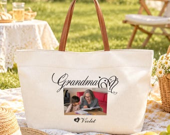 May include: A cream-colored canvas tote bag with brown faux leather handles. The front features a printed photograph of an adult and a child reading a book together, with the black text "Grandma" above and "& Violet" below the image.