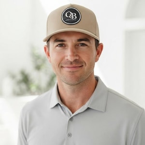 Könnte beinhalten: Eine beigefarbene Baseballkappe mit einem runden schwarzen Aufnäher auf der Vorderseite. Der Aufnäher zeigt den weißen Text C&B mit den Wörtern OATH & BRIN darunter. Die Kappe hat eine strukturierte Krone und einen gebogenen Schirm.